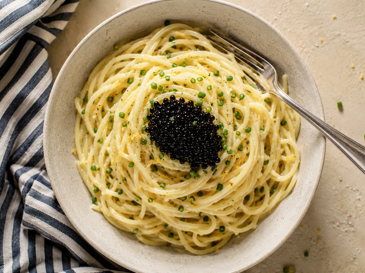 A bowl of buttery lemon spaghetti topped with a mound of black caviar and chives, a fork resting in the bowl.