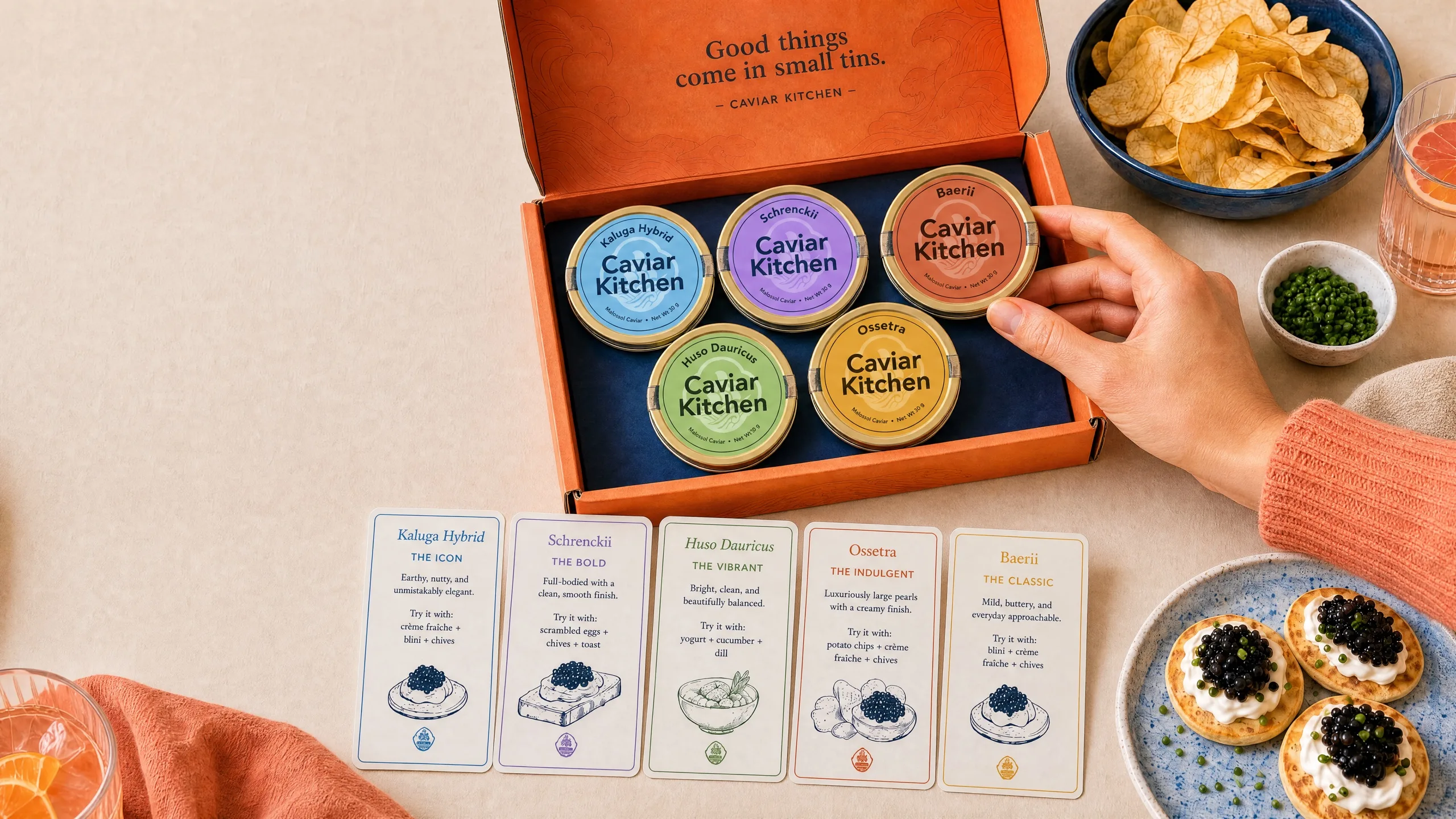 A Caviar Kitchen tasting box of five caviar tins with chips, blini, and drinks on a table.